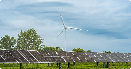 Solar panels in a grassy field with a wind turbine in the background under a blue, cloud-filled sky.