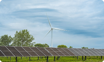 Solar panels in a grassy field with a wind turbine in the background under a blue, cloud-filled sky.